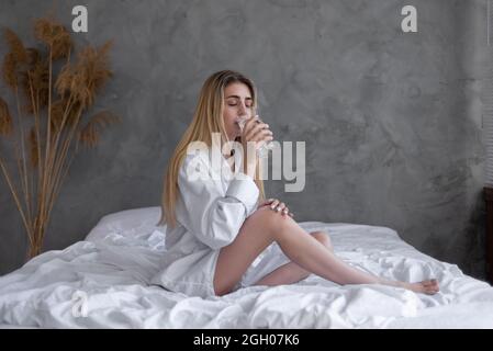 Un verre de plan propre dans la main d'une jeune femme. Femme en chemise blanche assise au lit, repas et boissons le soir du matin, Banque D'Images