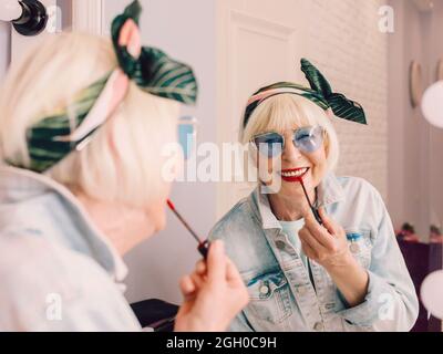 Femme âgée souriante et élégante en lunettes de soleil bleues et veste de Jean avec rouge à lèvres près du miroir dans un intérieur loft élégant.Style, mode, ma Banque D'Images