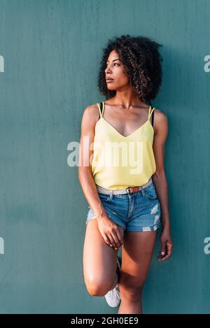 portrait vertical d'une femme afro-américaine avec un fond de mur vert. Elle s'habille d'un t-shirt jaune d'été et d'un Jean court et regarde d'un côté Banque D'Images