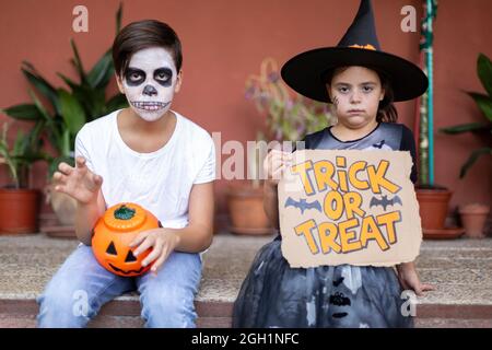 Garçon et fille en costumes typiques d'Halloween assis à l'entrée de leur maison. Banque D'Images