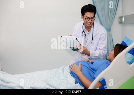 Homme médecin en robe blanche uniforme tenant le dossier et parlant à une femme enceinte se posant sur le lit dans la chambre d'hôpital avec des soins et heureux. Banque D'Images