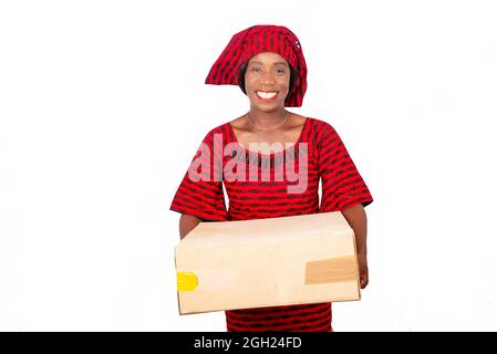 belle femme africaine mûre debout et vêtue d'un costume rouge en toile de longe tenant une boîte en carton dans la main tout en étant heureux Banque D'Images