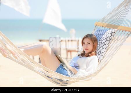 Femme posant sur le tissu balançant sur la plage de sable et utilisant ordinateur portable de travail et regardant à la caméra avec visage heureux. Concept de freelance et Banque D'Images