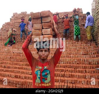 Un homme tient des briques sur sa tête après les avoir sortis des fours, tandis qu'elle traverse la rivière sur une planche pour les accueillir de l'autre côté à une briqueterie, pour distribuer les briques pour les entreprises de construction en dehors de Dhaka. Les employés de Brick Field travaillent 7 jours par semaine et sont payés BDT 350 (4.37 $) par jour, les propriétaires de chaque usine de Brick emploient entre 100-200 travailleurs et les gardent travailler du lever au coucher du soleil pendant la saison sèche. Le 4 septembre 2021 à Dhaka, au Bangladesh. (Photo de Habibur Rahman / Eyepix Group) Banque D'Images