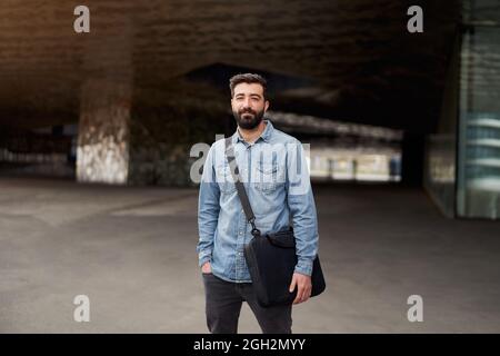 Homme d'affaires millénaire avec la barbe en plein air regarde la caméra sérieusement avec un regard intense. Portrait d'un homme en chemise décontractée avec sac pour ordinateur portable. Zoom avant Banque D'Images