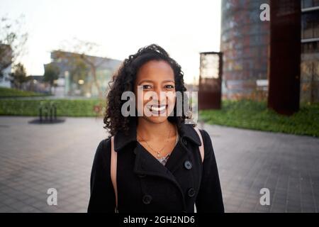Une femme d'affaires afro-américaine en sortie de travail sourit en regardant la caméra. La jeune femme se trouve à côté d'un immeuble de bureaux moderne. Banque D'Images