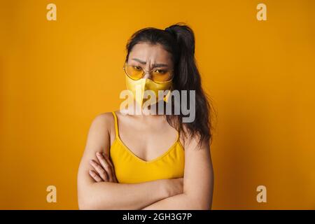 Frowning jeune asiatique décontracté femme portant un masque médical de protection debout sur fond jaune bras pliés Banque D'Images