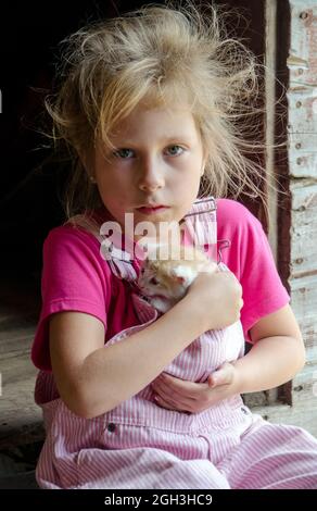 Une adorable petite fille de ferme en combinaison rose enfile un petit chaton à sa poitrine comme elle s'assoit sur un pas à l'extérieur de la grange Banque D'Images