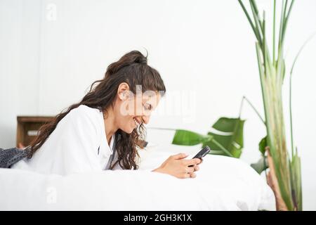 Une jeune femme hispanique souriante utilise un smartphone allongé sur le lit. Les utilisateurs des réseaux sociaux et les discussions en ligne grâce à la technologie. Banque D'Images