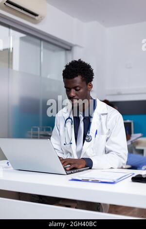 Portrait d'un médecin afro-américain assis au bureau à l'aide d'un ordinateur portable pour examen dans un cabinet médical à la clinique de santé. Black man avec périphérique moderne et fichiers de document sur table Banque D'Images