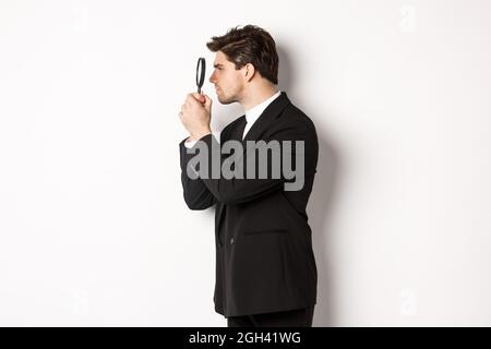 Photo de profil d'homme d'affaires beau en costume noir, regardant à travers la loupe et à la recherche de quelque chose, debout sur fond blanc Banque D'Images