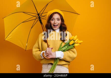 Jeune femme souriante aux cheveux rouges tenant des tulipes debout sous un parapluie sur fond de mur jaune Banque D'Images