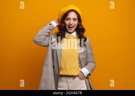 Bonne jeune femme avec de longs cheveux rouges dans le manteau et beret debout sur fond de mur jaune Banque D'Images
