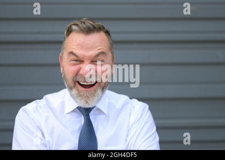 Homme d'affaires tirant un visage drôle en vissant ses yeux avec une expression d'angoisse tout en se tenant devant un mur extérieur gris Banque D'Images