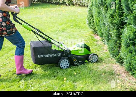 Une femme adulte non identifiable qui coupe l'herbe et tond une pelouse verte avec une tondeuse sans fil Greenworks 40v fonctionnant sur batterie. Banque D'Images