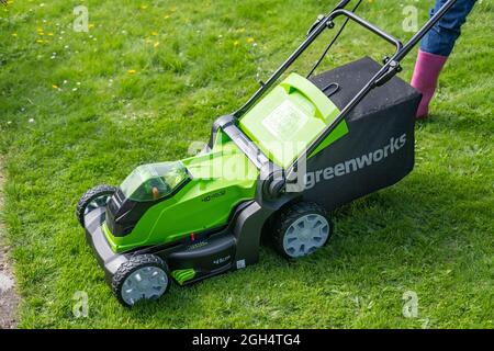 Une femme adulte non identifiable qui coupe l'herbe et tond une pelouse verte avec une tondeuse sans fil Greenworks 40v fonctionnant sur batterie. Banque D'Images