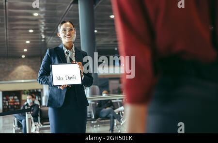 Femme portant un masque de protection debout au terminal de l'aéroport et portant une plaque d'identification pour recevoir un voyageur. Chauffeur de sexe féminin avec une pancarte sur le terminal des arrivées. Banque D'Images