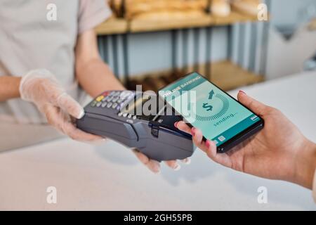 Gros plan sur le terminal de détention des employés de boulangerie gantés tout en obtenant le paiement du client à l'aide du système bancaire en ligne sur le téléphone Banque D'Images