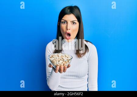 Jeune femme hispanique tenant des graines de citrouille bol effrayée et stupéfaite avec la bouche ouverte pour la surprise, incrédulité visage Banque D'Images