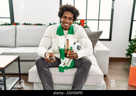 Un jeune homme de hooligan afro-américain boit de la bière en soutien à l'équipe de football assis sur le canapé à la maison. Banque D'Images