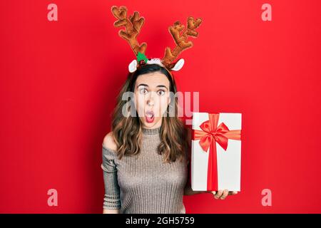 Jeune fille hispanique portant le chapeau de noël cerf tenant le cadeau effrayée et stupéfaite avec la bouche ouverte pour la surprise, le visage incrédulé Banque D'Images