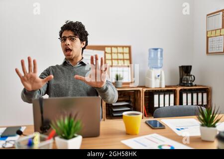 Jeune homme hispanique portant le style d'affaires assis sur le bureau peur et terrifié avec l'expression de peur stop geste avec les mains, criant en sho Banque D'Images