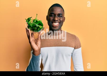 Jeune afro-américain tenant un bol avec de petits poivrons verts, en position positive et heureuse, souriant avec un sourire confiant montrant des dents Banque D'Images