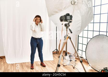 Femme hispanique d'âge moyen posant comme modèle dans le studio de photographie souffrant de maux de tête désespérés et stressés parce que la douleur et la migraine. Mains sur la hea Banque D'Images