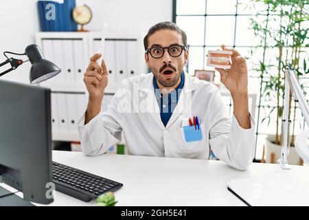 Jeune dentiste hispanique, homme tenant une prothèse dentaire et une brosse à dents effrayés et choqués par l'expression surprise et stupéfaite, la peur et le visage excité. Banque D'Images