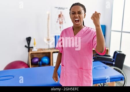 Une jeune femme afro-américaine travaillant à la clinique de rétablissement de la douleur en colère et folle levant le poing frustré et furieux tout en criant avec colère. Rage et agg Banque D'Images