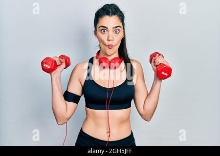 Jeune fille hispanique portant des vêtements de sport en utilisant des haltères faisant le visage de poisson avec la bouche et les yeux de accroupi, fou et comique. Banque D'Images