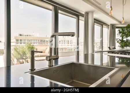 Détail du design intérieur de la cuisine moderne avec robinet et évier chromés installés dans un comptoir brillant devant une grande fenêtre donnant sur le paysage urbain Banque D'Images