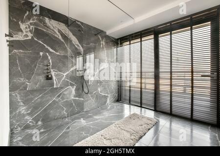 Intérieur d'une salle de bain spacieuse et contemporaine avec douche sur un mur en marbre et une cloison en verre près de la fenêtre avec volets dans un appartement de luxe moderne Banque D'Images