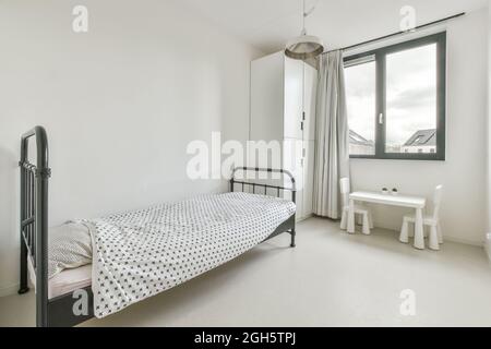 Décoration intérieure simple et minimaliste de la chambre blanche avec lit simple et armoire placée près de la fenêtre et petite table avec chaises dans l'appartement moderne Banque D'Images