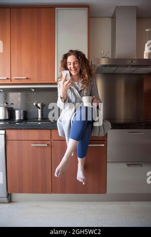 Femme d'affaires aux cheveux bouclés assise dans la cuisine, prenant une infusion tout en utilisant son smartphone et en travaillant à la maison Banque D'Images