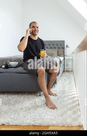 Homme joyeux assis sur le lit et parlant sur le téléphone portable pendant le petit déjeuner le matin à la maison Banque D'Images