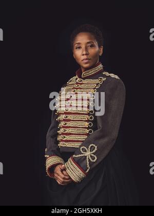 Femme adulte afro-américaine confiante dans une veste élégante regardant l'appareil photo dans un studio sombre sur fond noir Banque D'Images