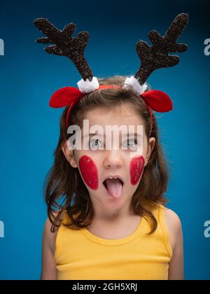 Surprise fille avec des joues peintes en rouge portant des cornes et des oreilles de cerf jouet et regardant la caméra sur fond bleu pendant qu'elle colle sa langue Banque D'Images