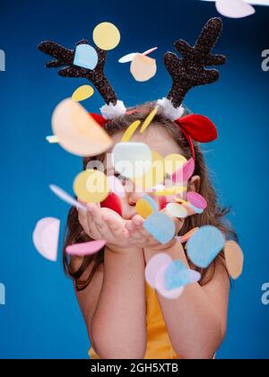 fille aux joues peintes en rouge et au bandeau antinet qui soufflait de confettis de papier vif tout en se tenant sur fond bleu Banque D'Images