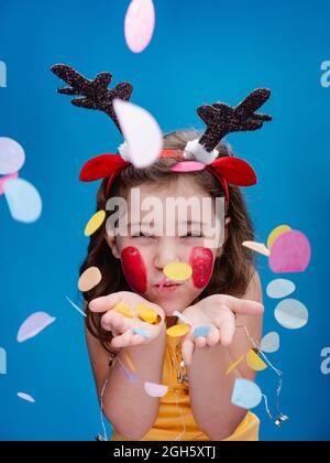 fille aux joues peintes en rouge et au bandeau antinet qui soufflait de confettis de papier vif tout en se tenant sur fond bleu Banque D'Images