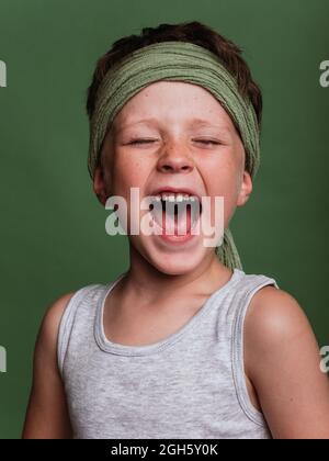 Jeune garçon de karaté positif dans le foulard hachimaki criant bruyamment avec les yeux fermés dans le studio sur fond vert Banque D'Images