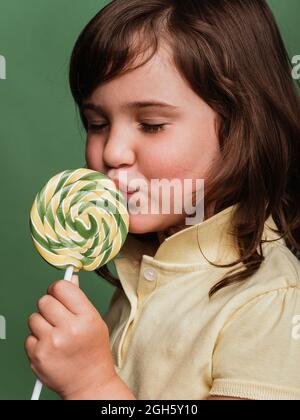 Drôle de préteen enfant léchant doux tourbillon lolipop sur fond vert en studio avec les yeux fermés Banque D'Images