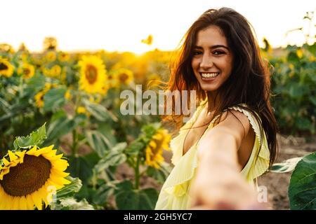 Vue latérale d'une jeune femme hispanique à cheveux longs et gaie vêque de couleur jaune, regardant l'appareil photo et la main en plein air comme une invitation à la suivre dans une infinité Banque D'Images