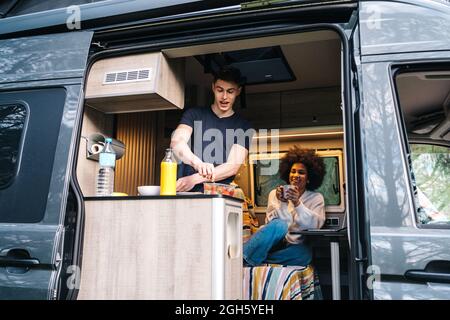 Un jeune homme prépare un petit déjeuner sain tandis qu'une petite amie afro-américaine utilise des gadgets pendant le voyage d'été ensemble dans une camionnette de camping Banque D'Images