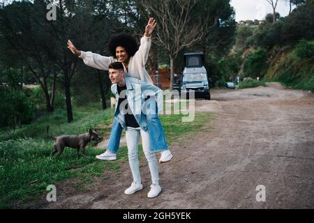 Jeune homme offrant une petite amie ethnique joyeuse en train de faire une promenade en pickyback tout en regardant loin contre le campeur et Thai Ridgeback sur la voie Banque D'Images