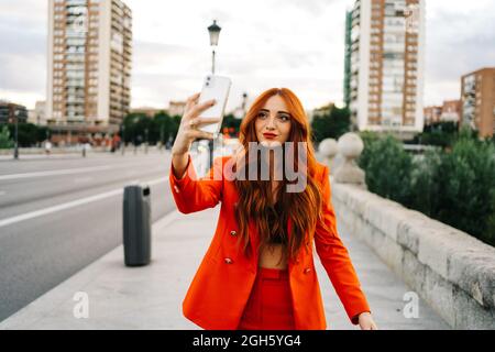 Femme ravie avec cheveux rouges et en costume orange marchant dans la rue en ville et prenant autoportrait sur smartphone Banque D'Images