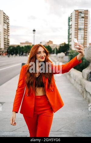 Femme ravie avec cheveux rouges et en costume orange marchant dans la rue en ville et prenant autoportrait sur smartphone Banque D'Images