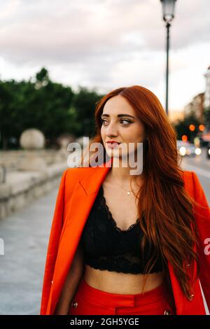 Charmante femme avec de longs cheveux rouges et dans un costume orange tendance se tenant dans la rue le soir et regardant loin Banque D'Images