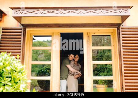 Vue de la terrasse de couple multiracial embrassant tout en se tenant dans la chambre près des portes de la maison en bois et souriant Banque D'Images