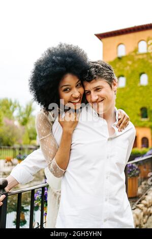 Femme noire affectueuse avec une coiffure afro embrassant l'homme souriant de derrière tout en se tenant sur le pont dans le parc en été et en appréciant le week-end ensemble Banque D'Images